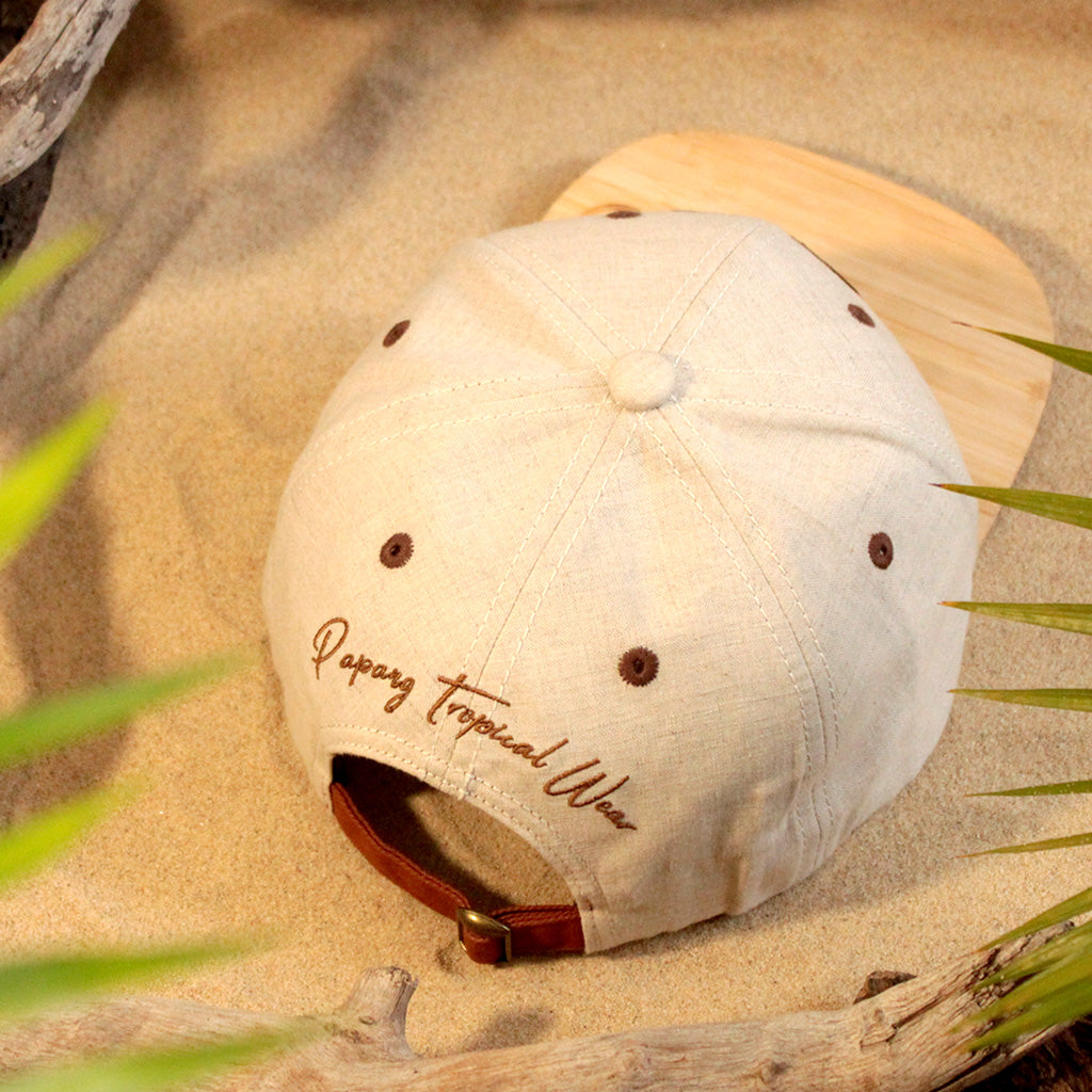 Vue arrière d'une casquette de marque Papang Tropical Wear de couleur lin sablé. Elle possède une visière en bois de bambou gravé du logo de la marque. La casquette est déposée sur le sable d'une plage de l'île de La Réunion.