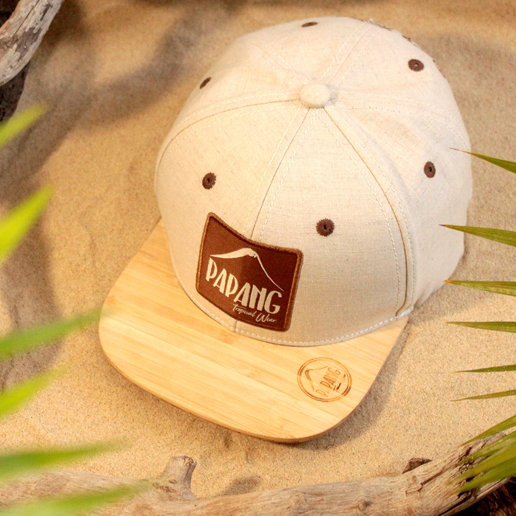 Vue avant d'une casquette Papang Tropical Wear de couleur lin sablé. La casquette possède une visière en bois de bambou gravé du logo de la marque. La casquette est posée sur une plage de l'île de La Réunion.