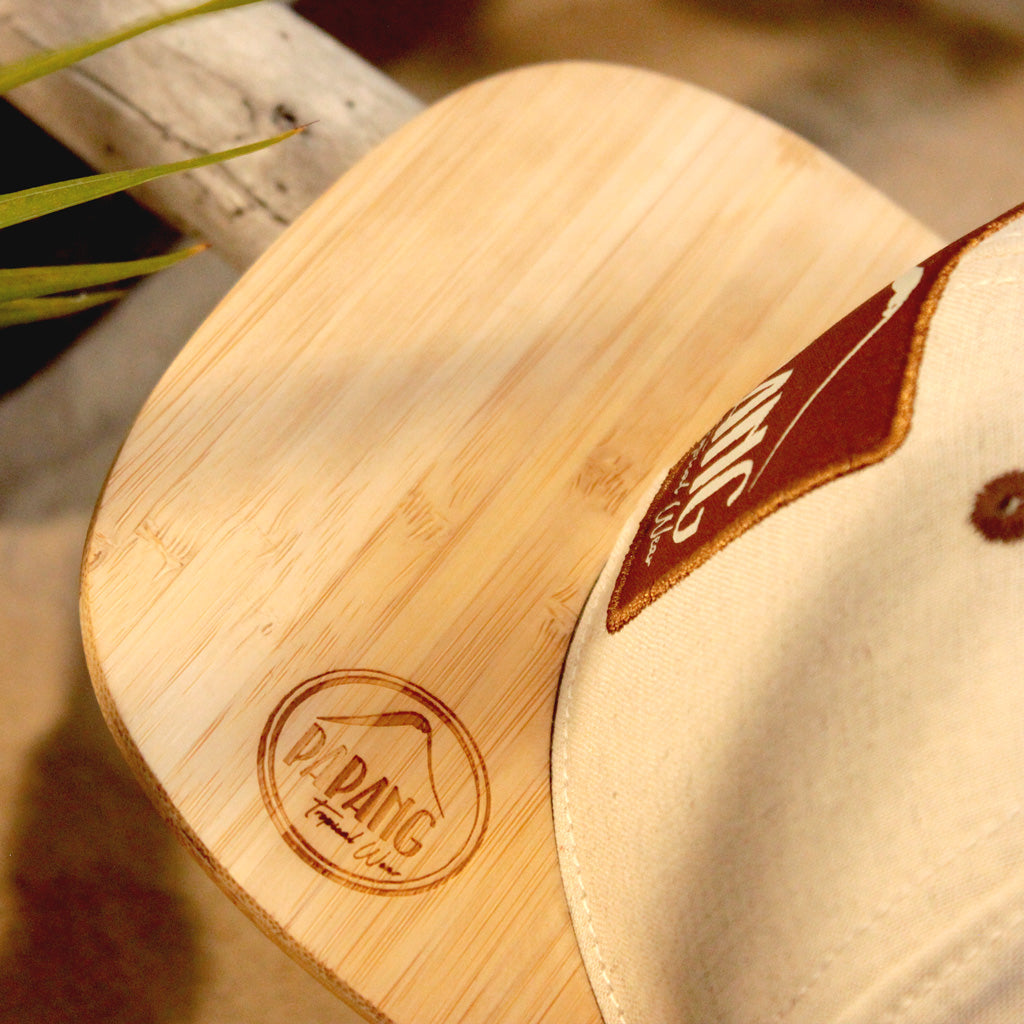Gros Plan d'une casquette de marque Papang Tropical Wear de couleur Lin sable. Elle possède une visière en bambou gravé du logo de la marque. La casquette est déposée sur une plage de l'île de La Réunion.